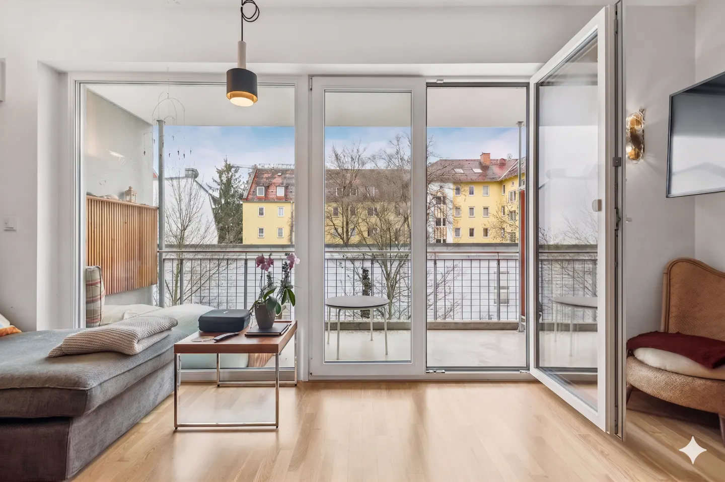 Bright living room with wood floors, gray sofa, and open balcony doors. Outside view of yellow buildings and trees.
