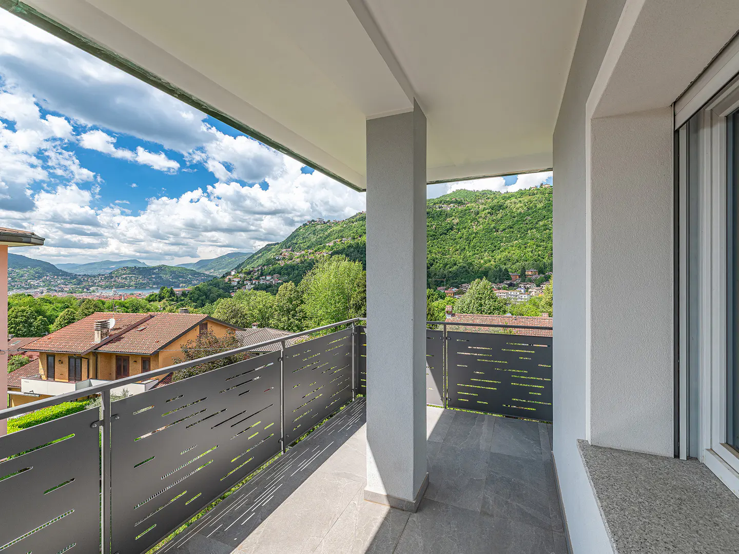 Balcony view: Gray metal railing, gray tile floor, white walls. Lush green hills and blue sky with clouds in the background.
