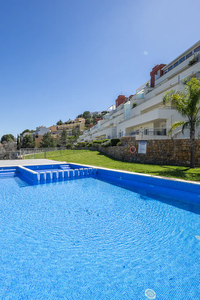 A blue tiled pool with steps, a grassy area, and white buildings on a hill under a clear blue sky.