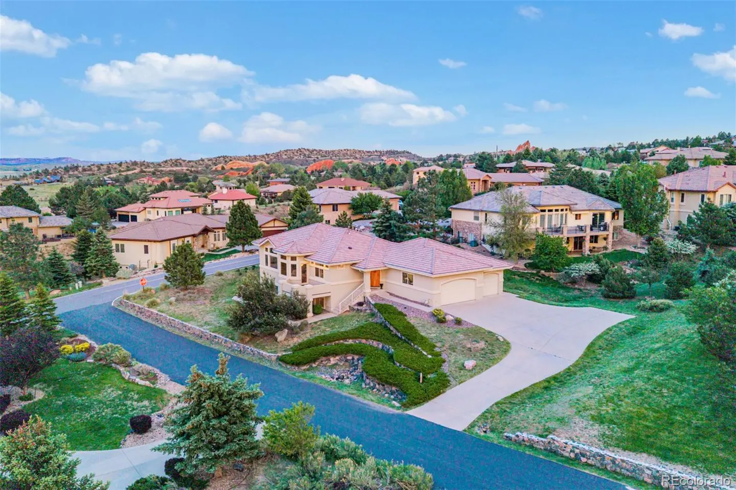 Aerial view of a beige house with a red tile roof, surrounded by green trees and other houses in a hilly neighborhood. Blue sky with clouds.