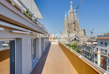 Balcony view of Sagrada Familia in Barcelona. The balcony has a wood floor and a beige wall with windows. The sky is blue.