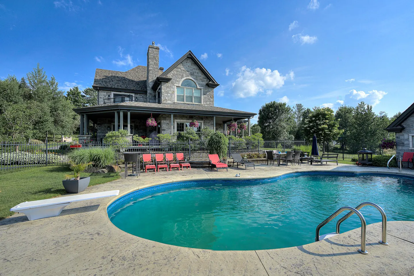 A stone house with a pool, diving board, and red lounge chairs sits under a blue sky with scattered clouds.