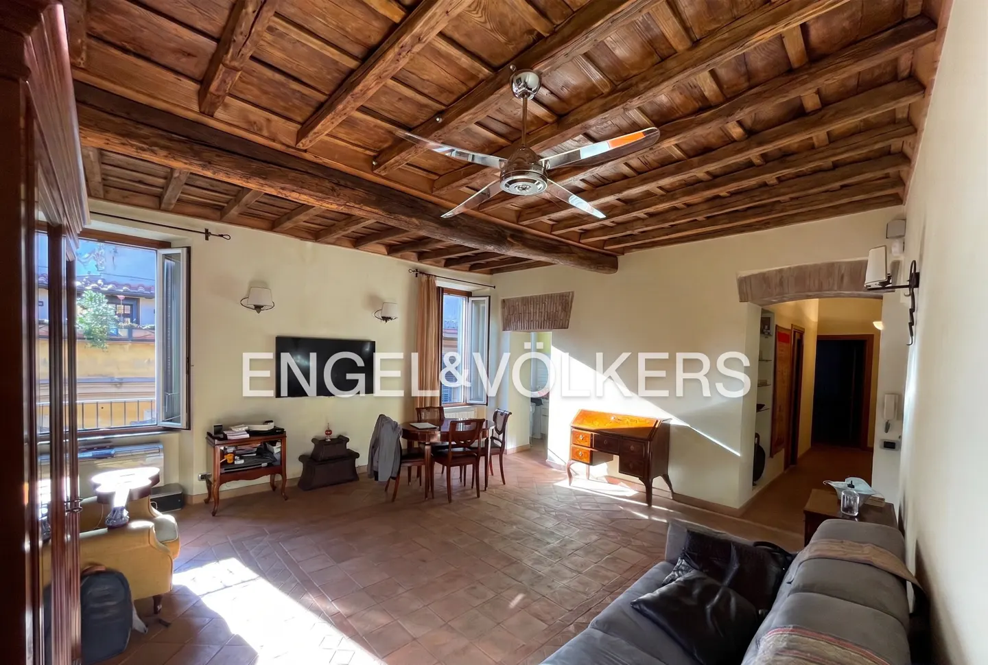 A spacious living room with a wooden beamed ceiling, terracotta tile floor, and a ceiling fan. There is a sofa, desk, and dining table.