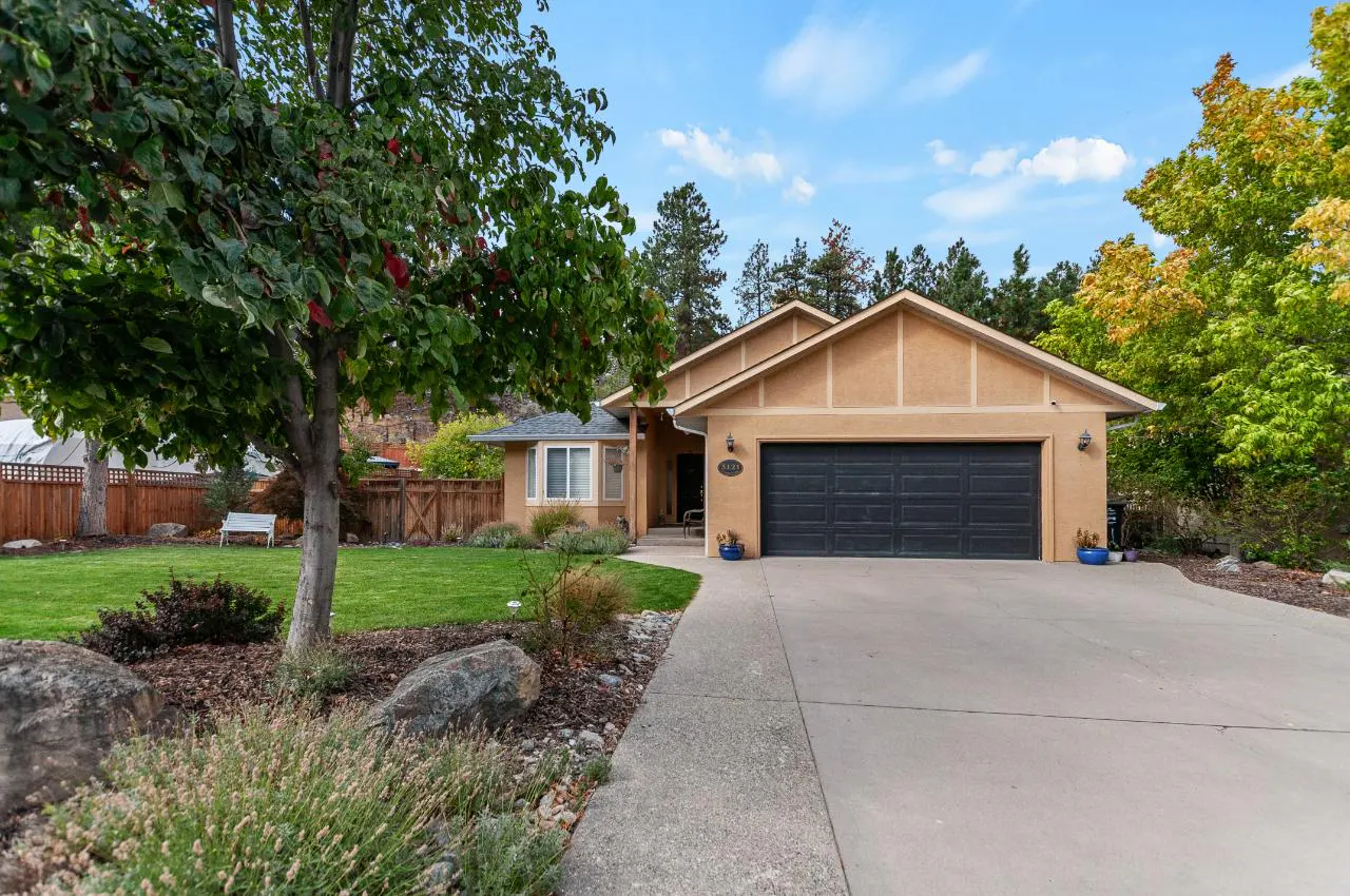 Tan single-story home with a dark gray garage door, green lawn, and trees under a blue sky.