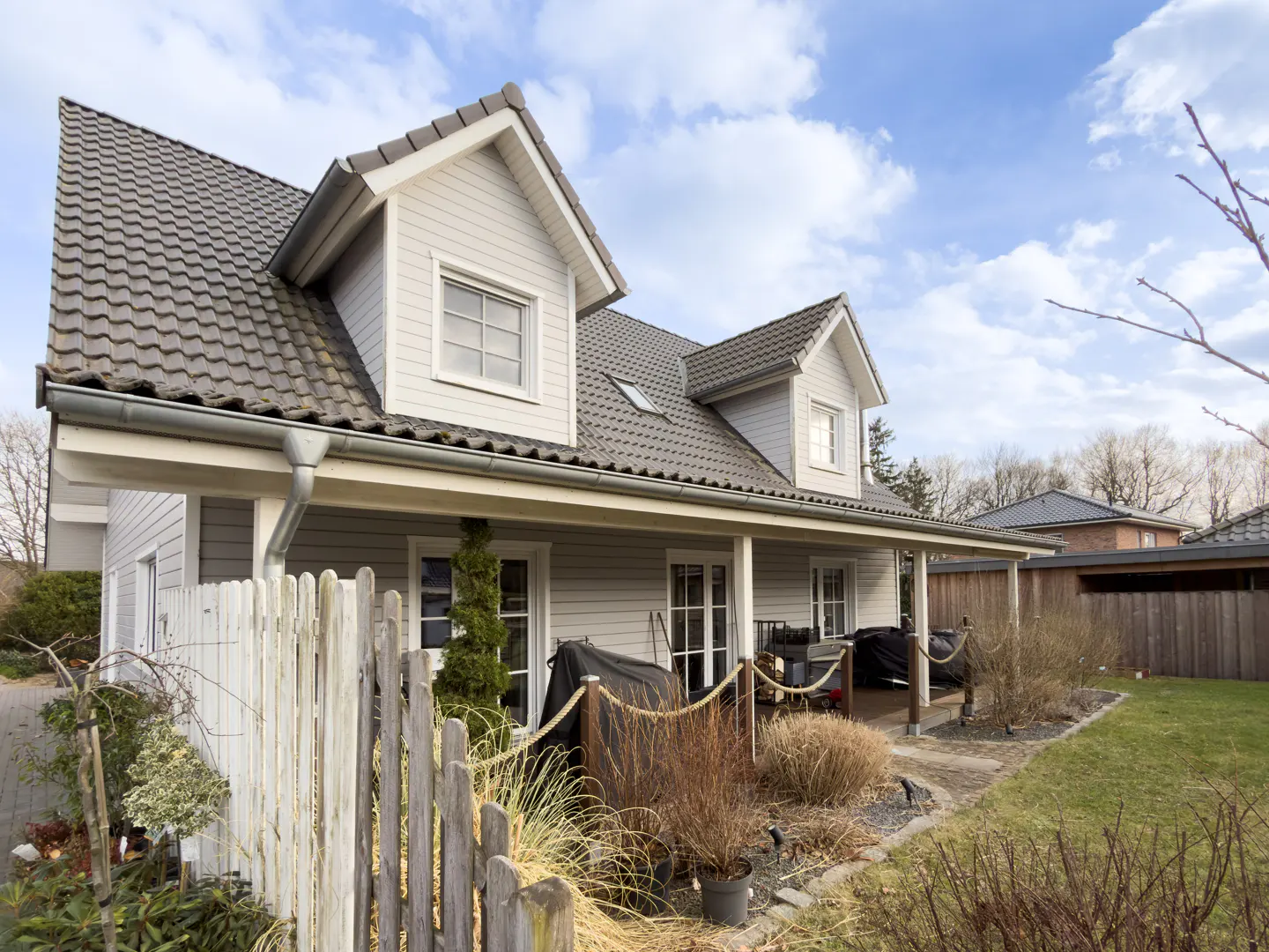 Exterior view of a two-story gray house with a dark gray roof, white trim, and a covered porch. A weathered white fence borders the front yard.
