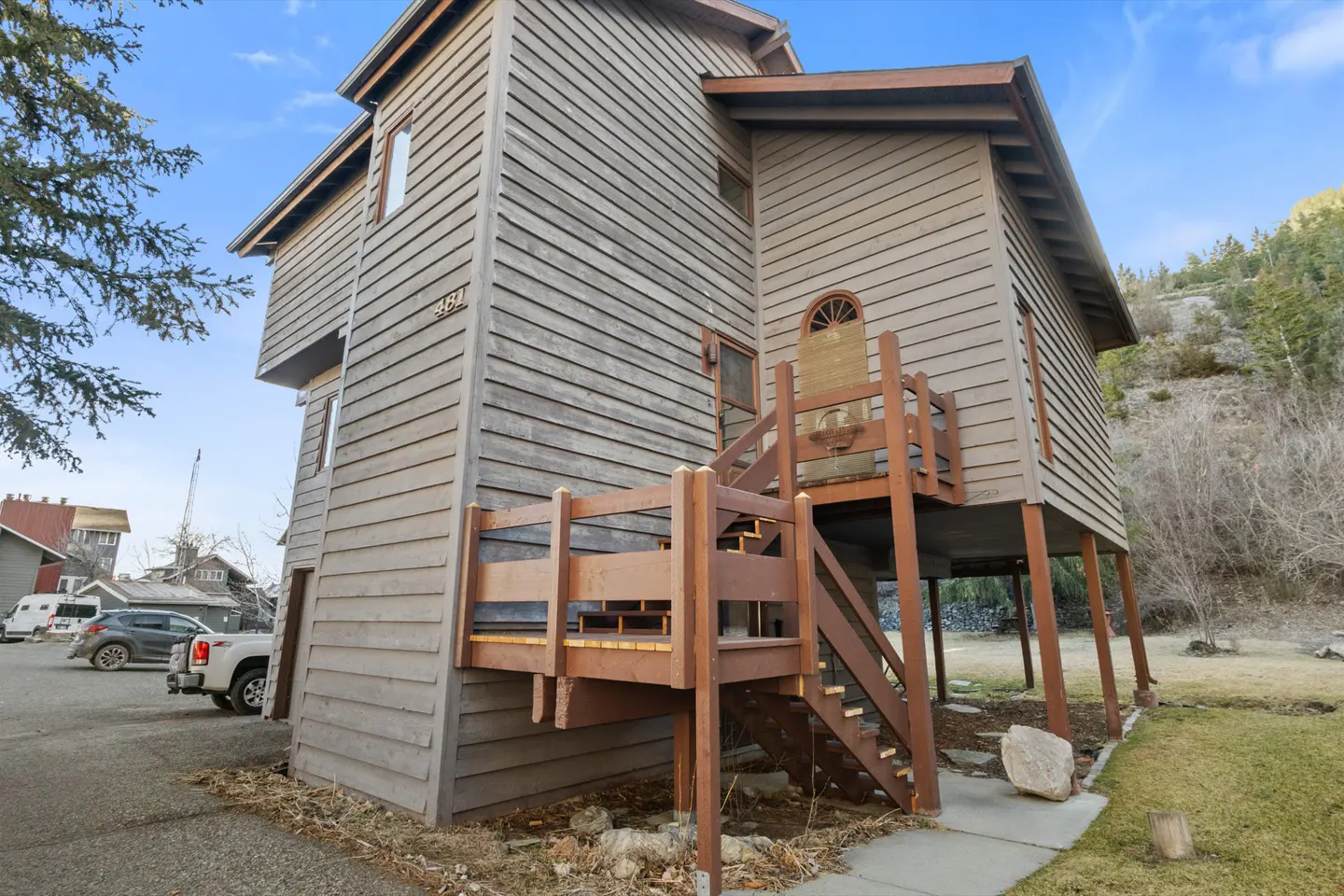 A three-story wood-sided house with a brown deck and stairs. The house is elevated on stilts, with a mountain in the background.