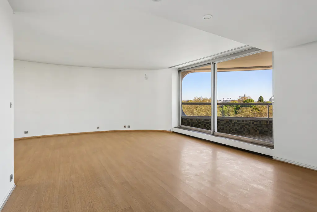 Empty room with light wood floors, white walls, and large sliding glass doors to a balcony with a view of trees and a blue sky.