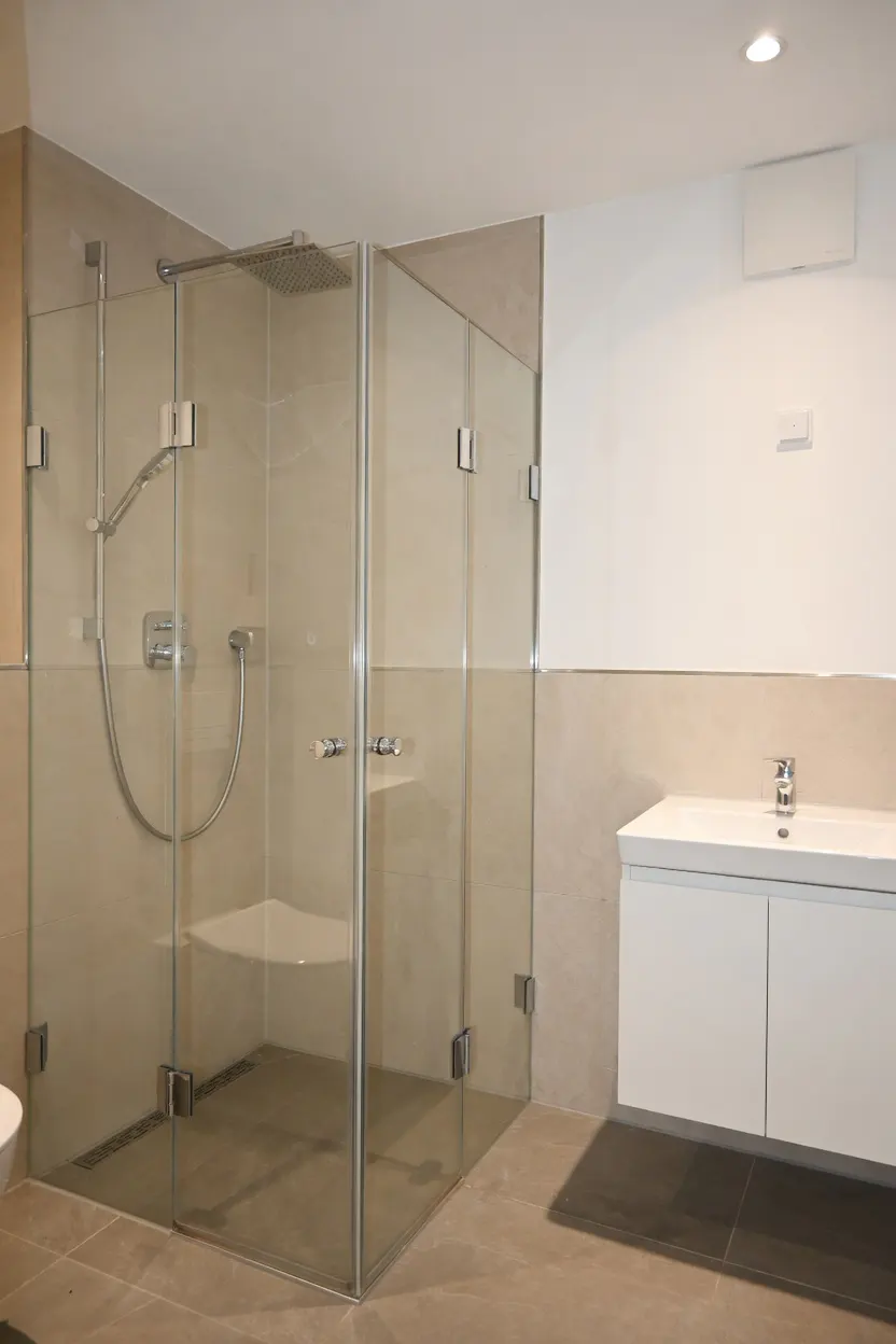 Modern bathroom with glass shower enclosure, white vanity, and beige tile. The shower has a built-in seat and rainfall showerhead.