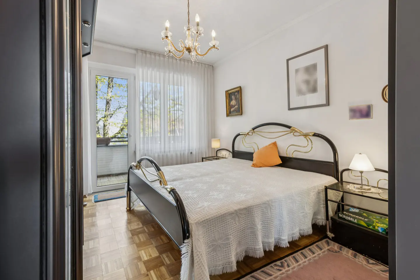 Bedroom with black metal bed frame, white bedding, orange pillow, and balcony view. Chandelier and parquet floor.