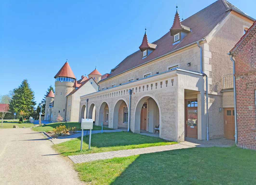 Exterior view of a beige castle-like building with red-roofed towers and arched walkways, set against a clear blue sky.
