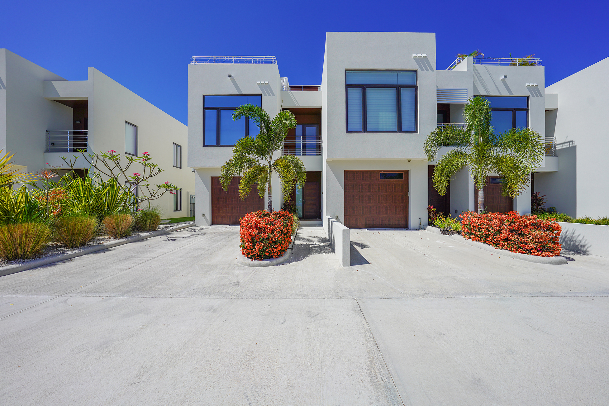 Modern white townhouses with brown garage doors under a clear blue sky. Palm trees and red flowering bushes add color to the landscape.