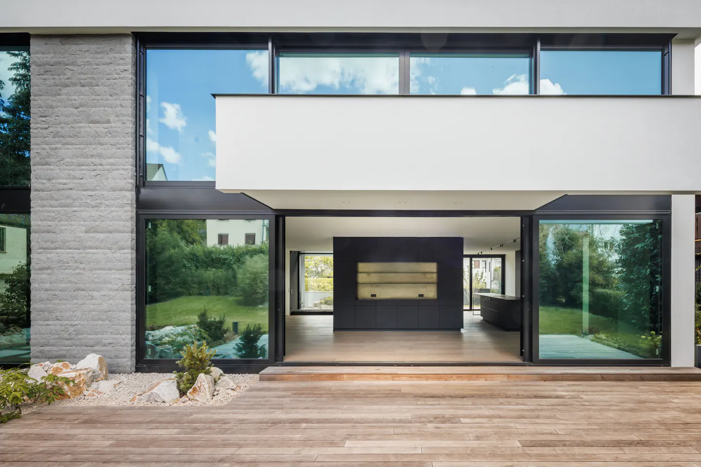 Modern home exterior with a wooden deck, large sliding glass doors, and a stone accent wall. The interior features a dark kitchen.