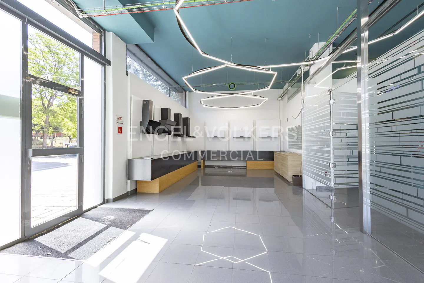 Empty commercial space with gray tile floors, a teal ceiling, and modern lighting fixtures. Glass-walled offices are on the right.