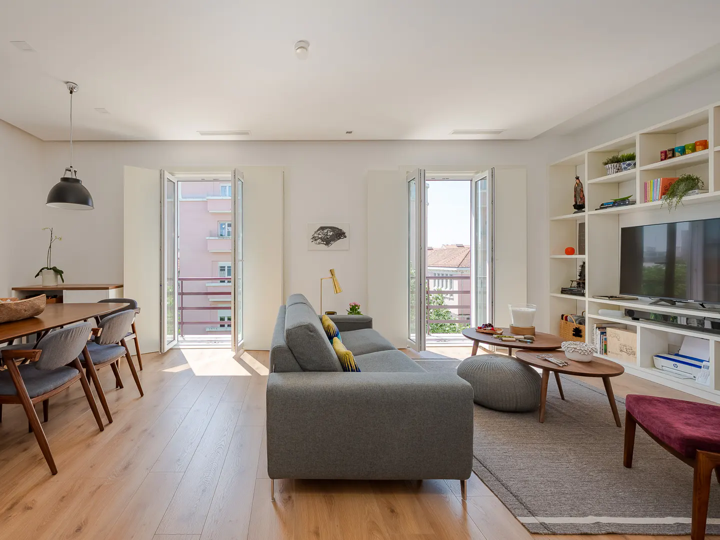 Bright living room with wood floors, gray sofa, dining table, and open balcony doors. A white bookshelf with a TV is on the right.