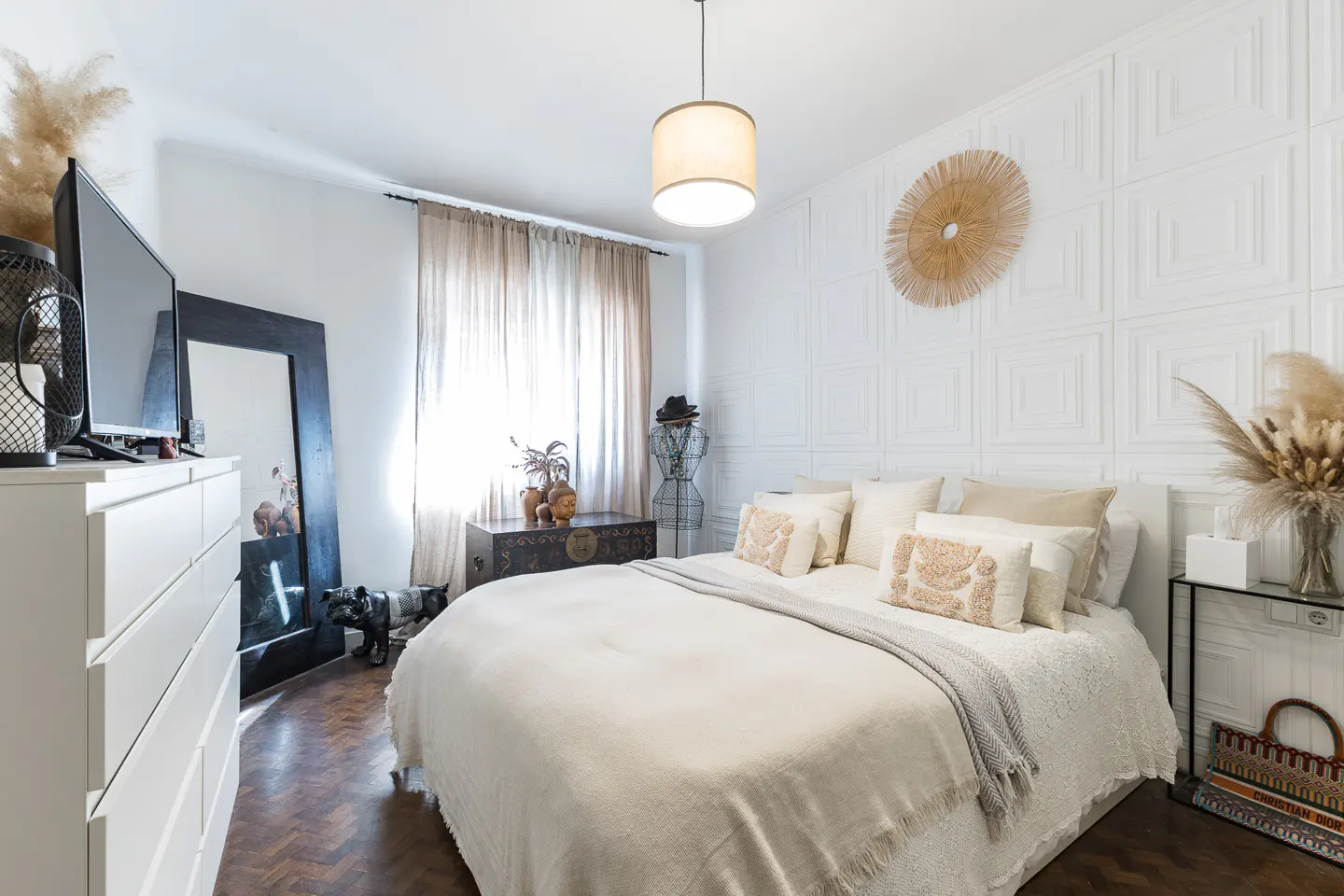Bright bedroom with white walls, textured headboard, and neutral bedding. A large mirror and dresser are on the left. A straw decoration hangs above the bed.