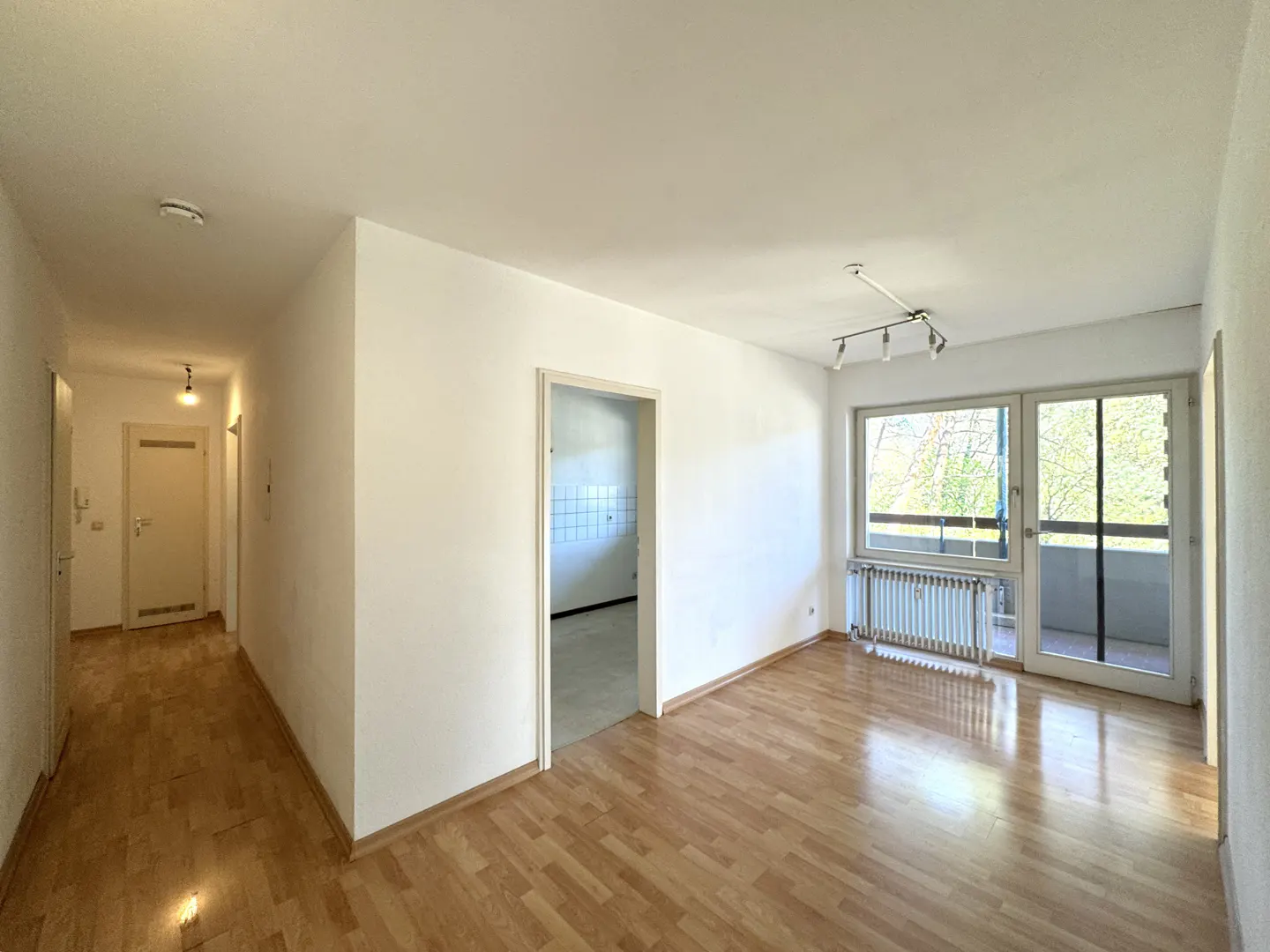 Empty apartment with wood floors, white walls, and a hallway leading to a door. A doorway leads to a kitchen. A window overlooks trees.