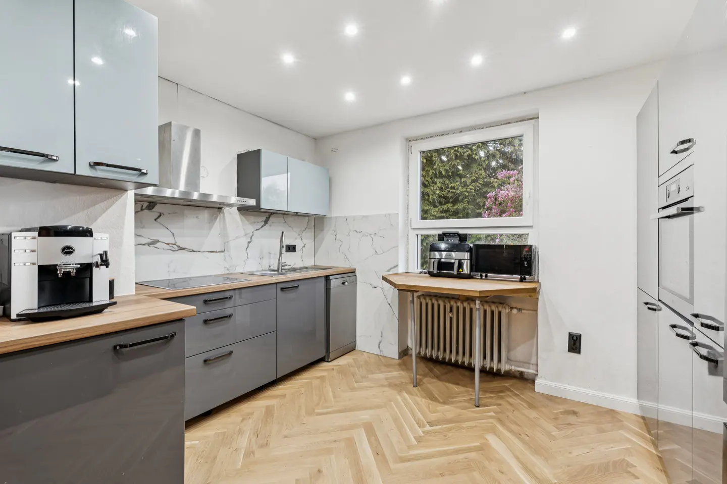 Bright kitchen with gray cabinets, wood countertops, and herringbone floors. A window overlooks trees. Appliances include a coffee maker and microwave.