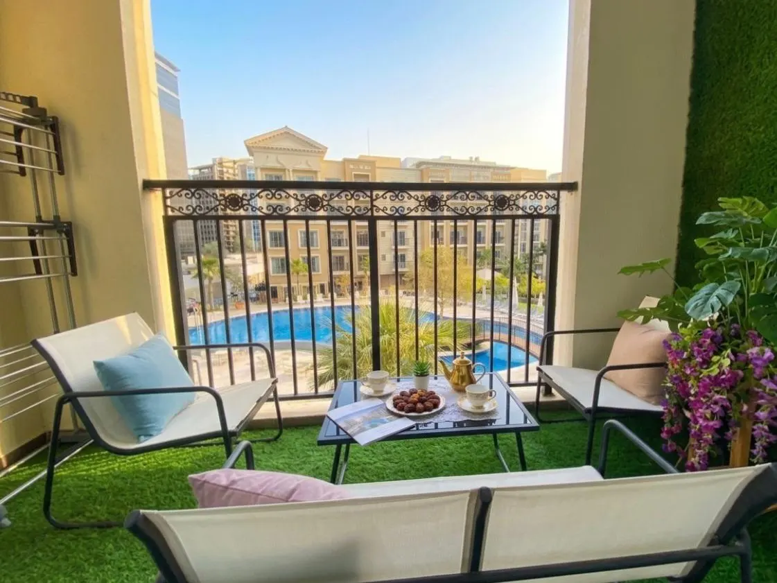 Balcony view with seating, table set with tea, and a pool view. Ornate railing and green turf create a relaxing outdoor space.