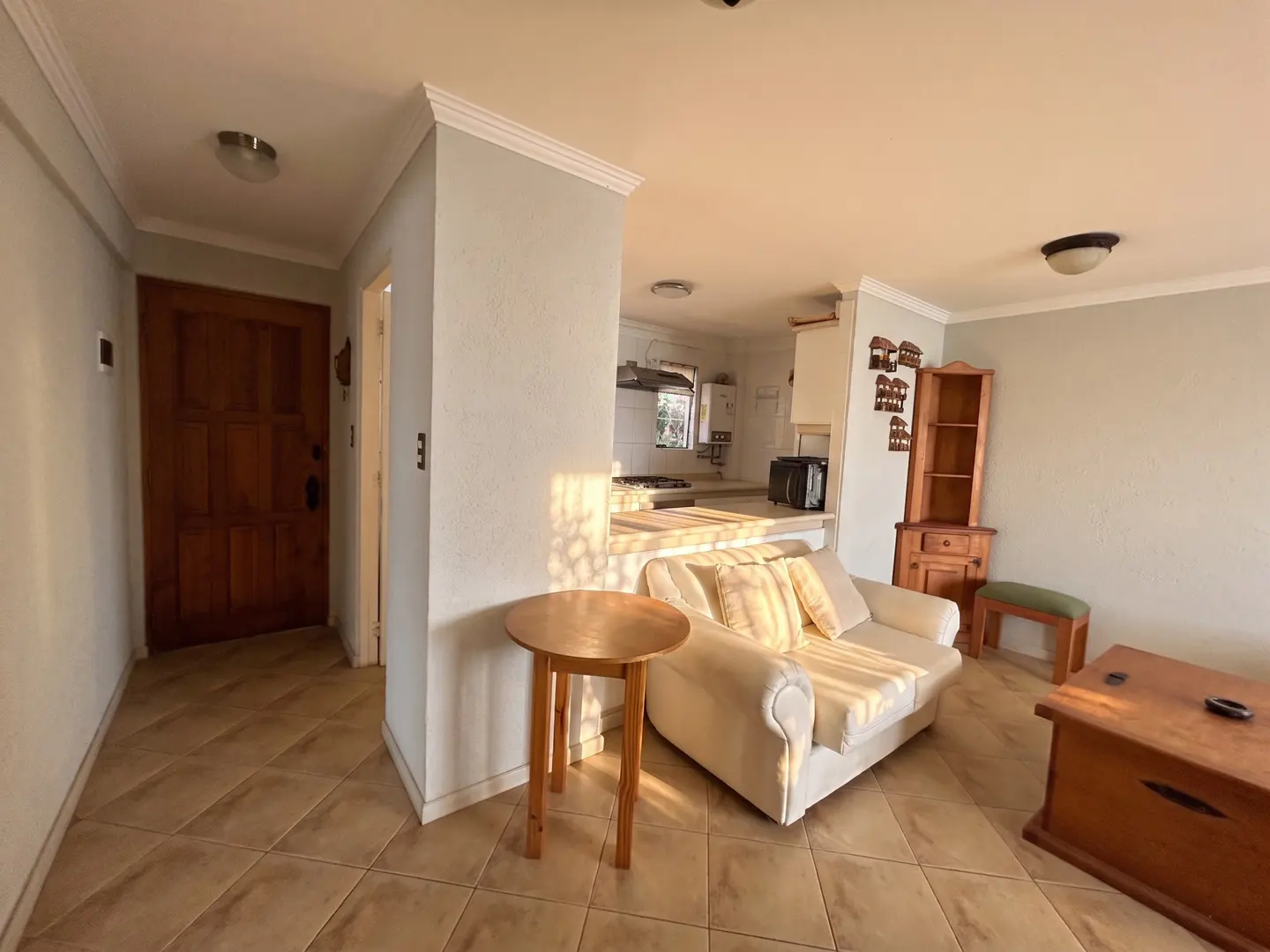 A bright living room with a cream sofa, wooden furniture, and a view into the kitchen. The floor is tiled in a diamond pattern.