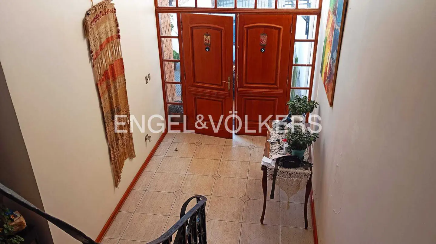 Entryway with open wood double doors, tiled floor, and a table with plants. A woven wall hanging is on the left.