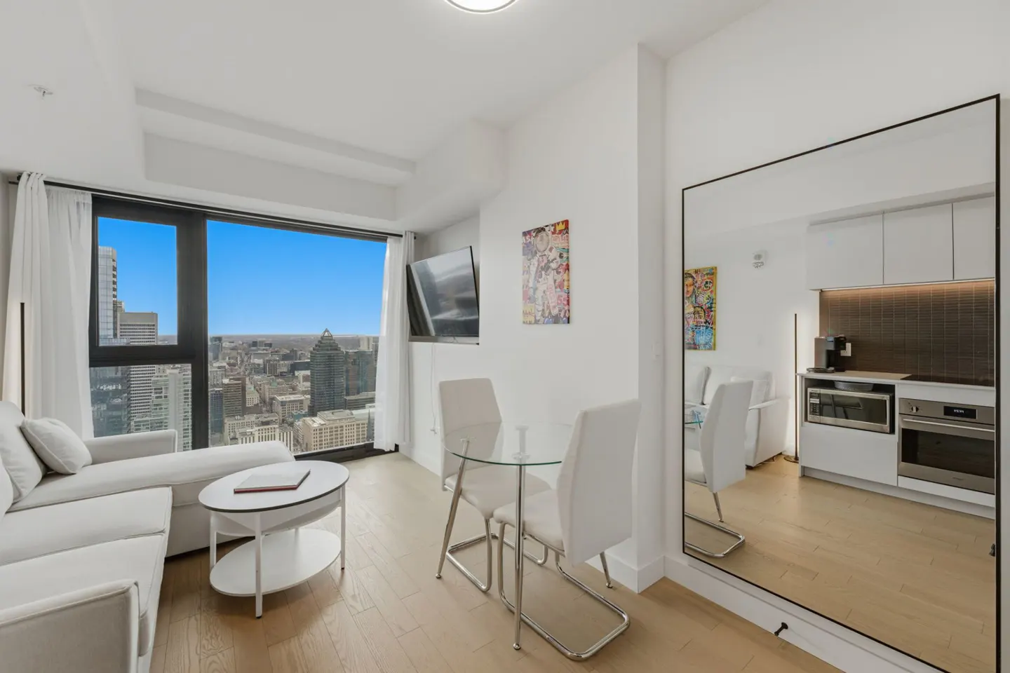 Bright condo interior with a white sofa, glass table, and city view from a large window. A mirror reflects the kitchen.