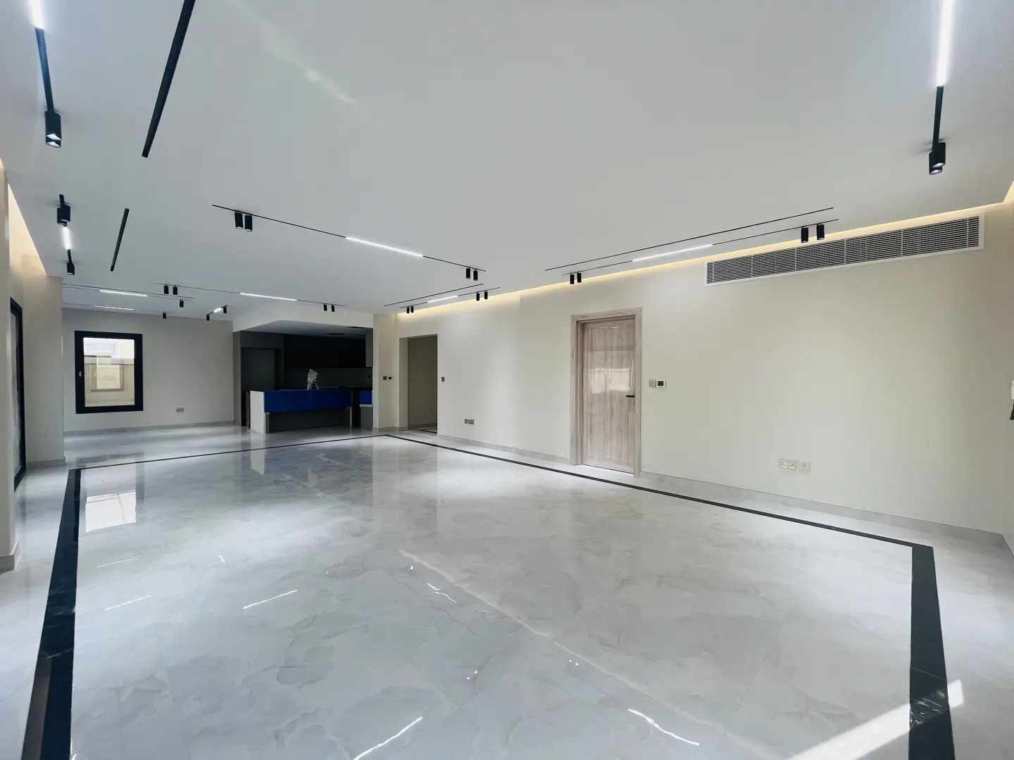 A bright, empty room with white walls, a light wood door, and shiny marble floors. Black track lighting is on the ceiling.