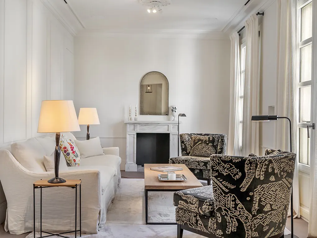 Bright living room with white walls, sofa, patterned chairs, and a marble fireplace with a mirror above. Natural light streams through the window.
