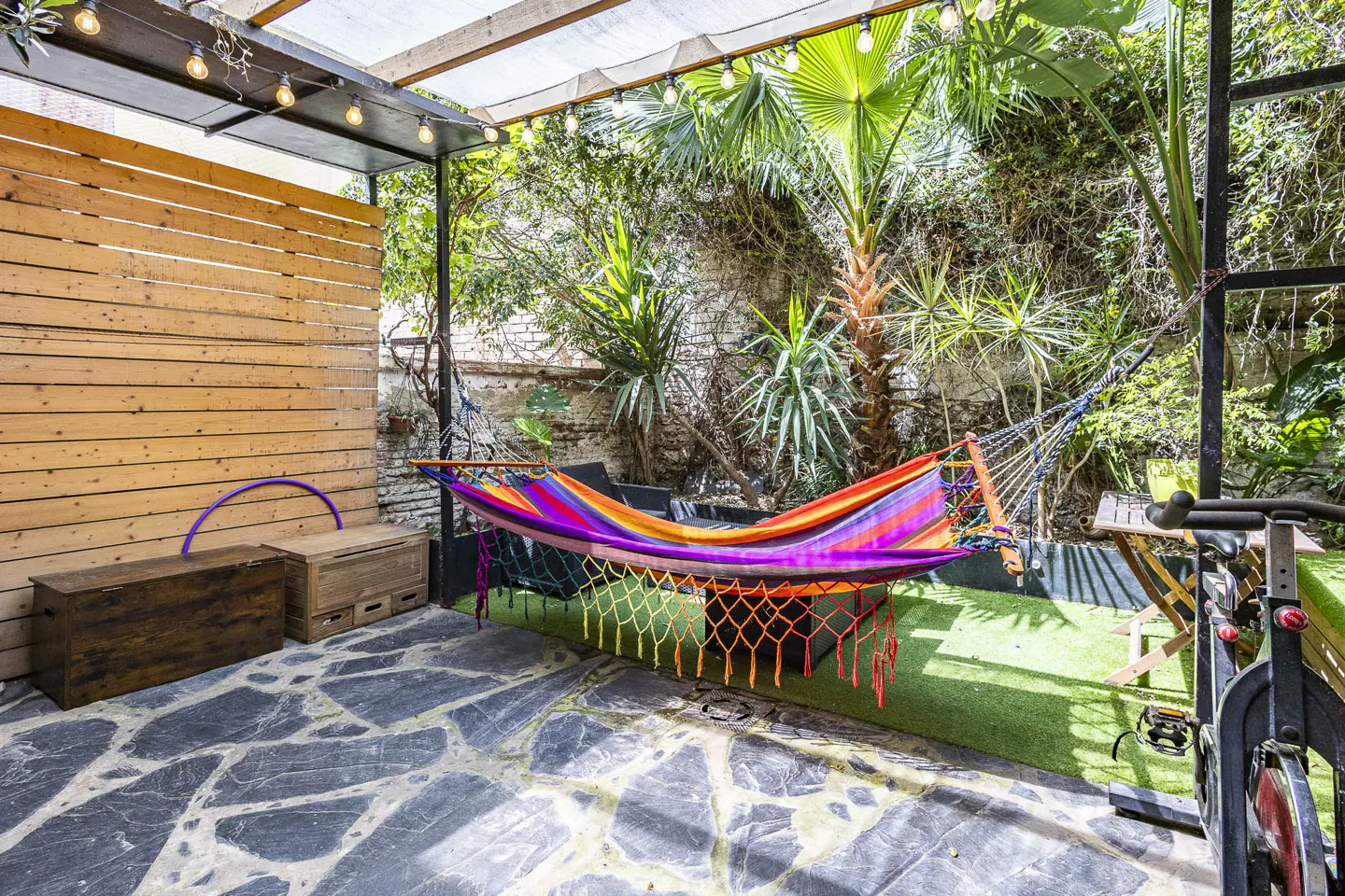 Outdoor patio with a colorful hammock, stone floor, wood wall, and lush greenery. String lights hang above.