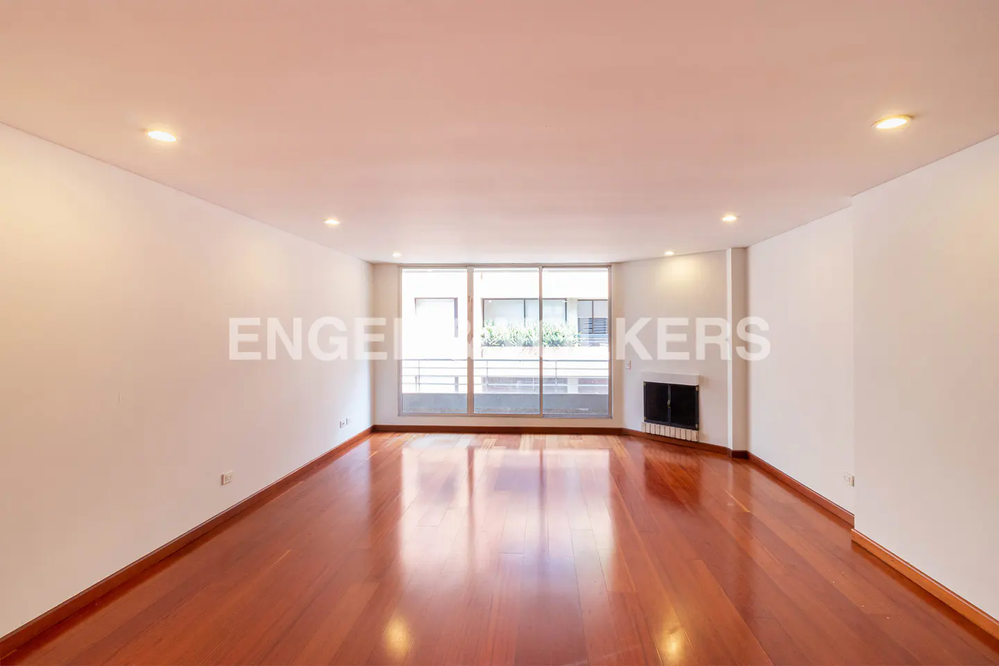 Empty living room with hardwood floors, white walls, and a large window overlooking a balcony with greenery.