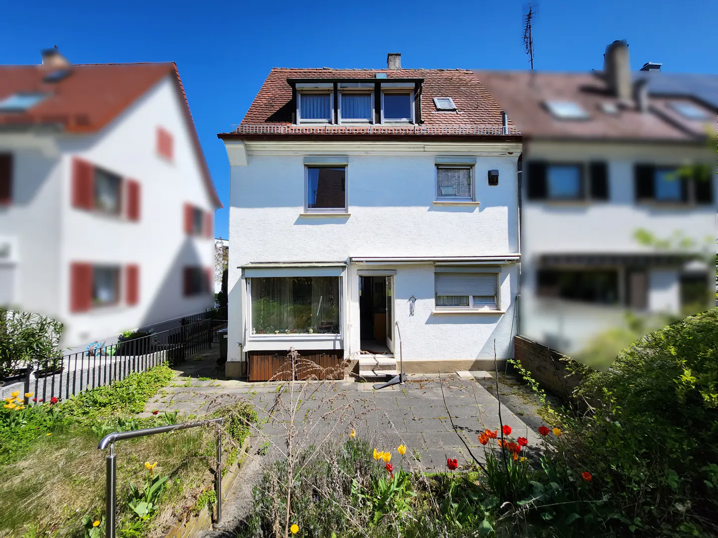 Two-story white house with a red-tiled roof, surrounded by greenery and flowers under a clear blue sky.