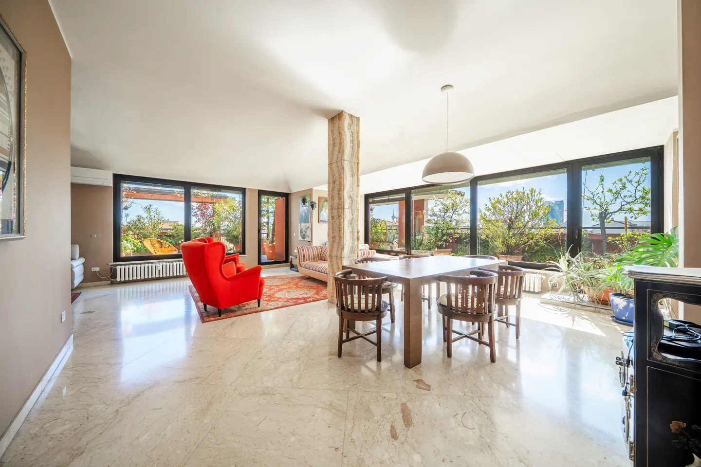 Bright, open living space with marble floors, a red armchair, and a wooden dining table set. Large windows offer views of greenery.