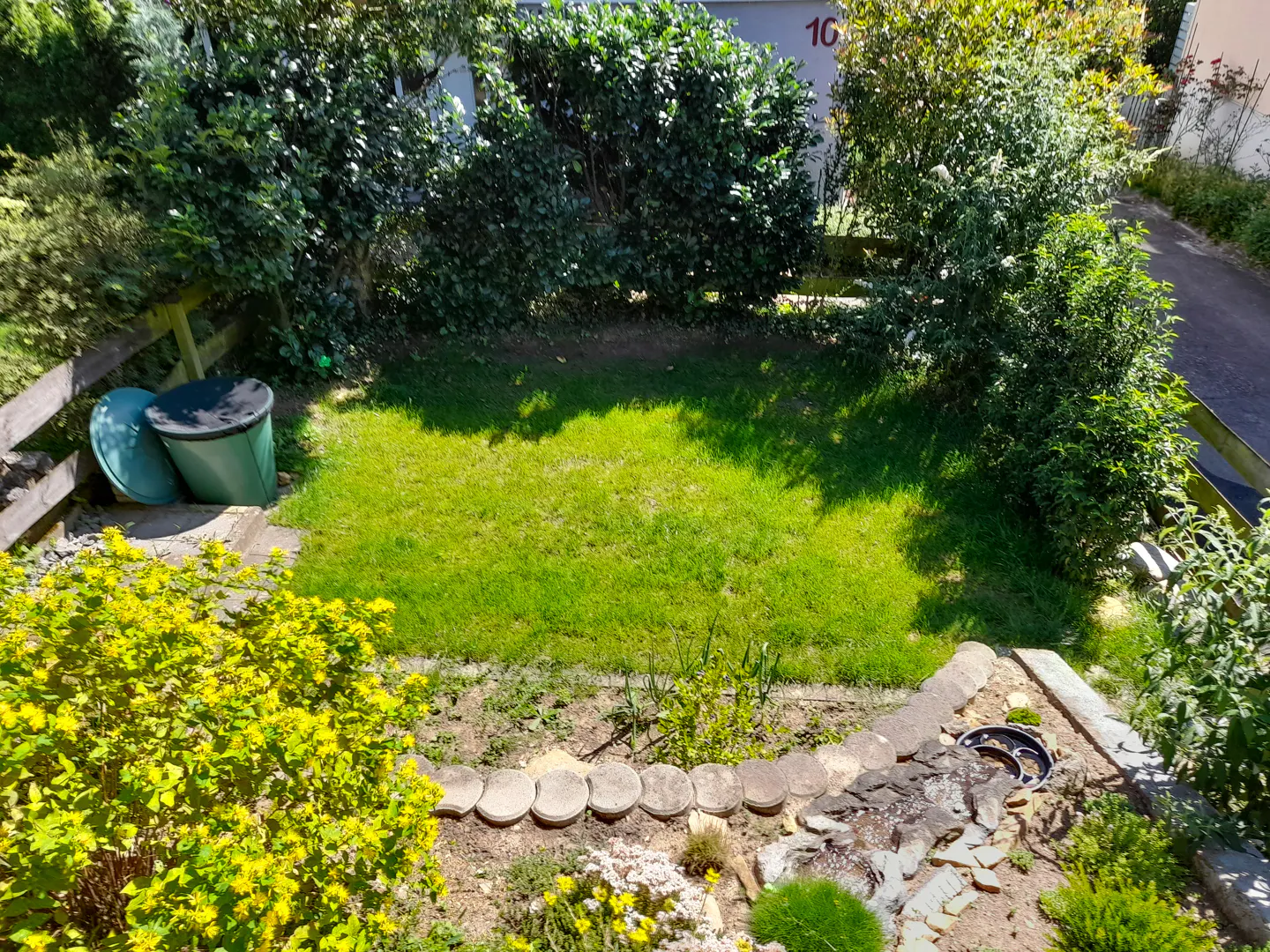 A small green lawn is surrounded by bushes and flowers. A green water barrel is on the left.