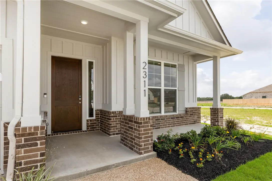 Front porch of a house with a brown door, white columns, brick accents, and the address "2311" displayed. A flower bed adds a touch of color.