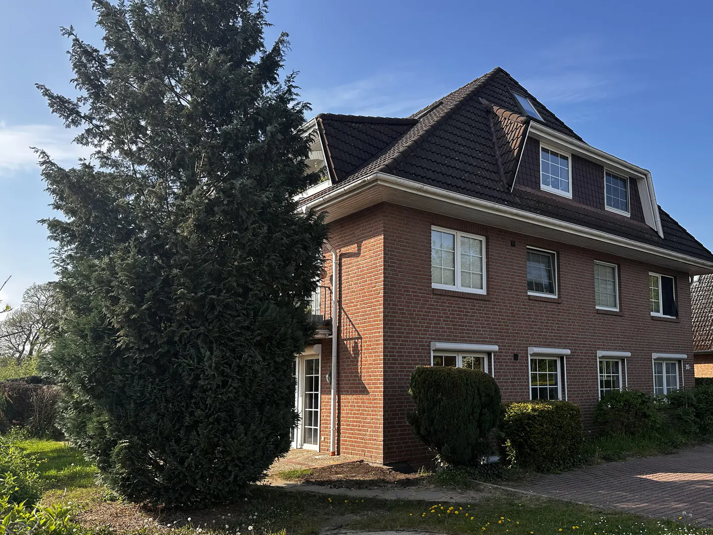 A two-story brick house with a dark roof and white-framed windows, partially obscured by a large evergreen tree.