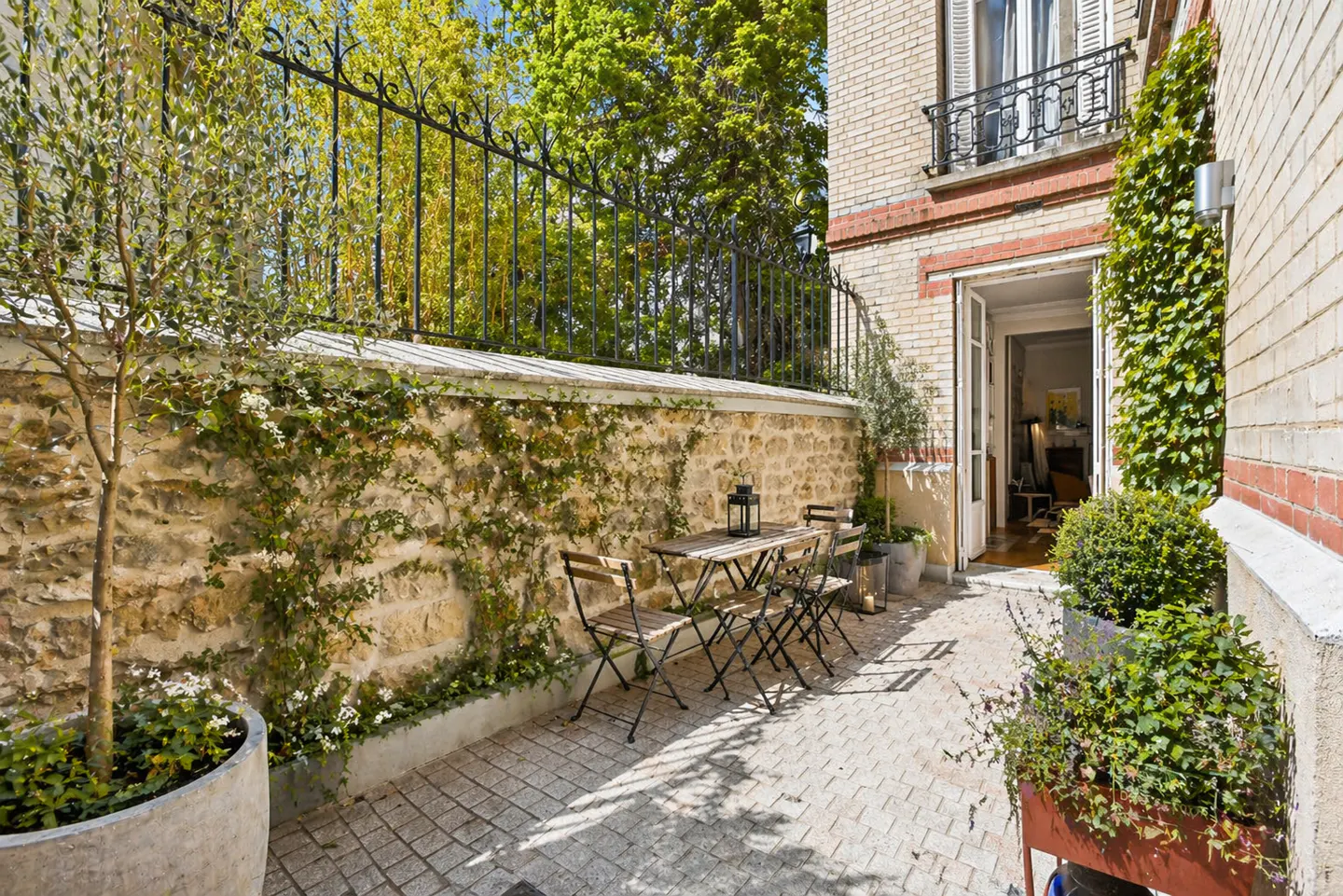 Outdoor patio with stone wall, iron fence, and brick building. Table and chairs sit on a stone-paved area, with greenery and an open doorway.