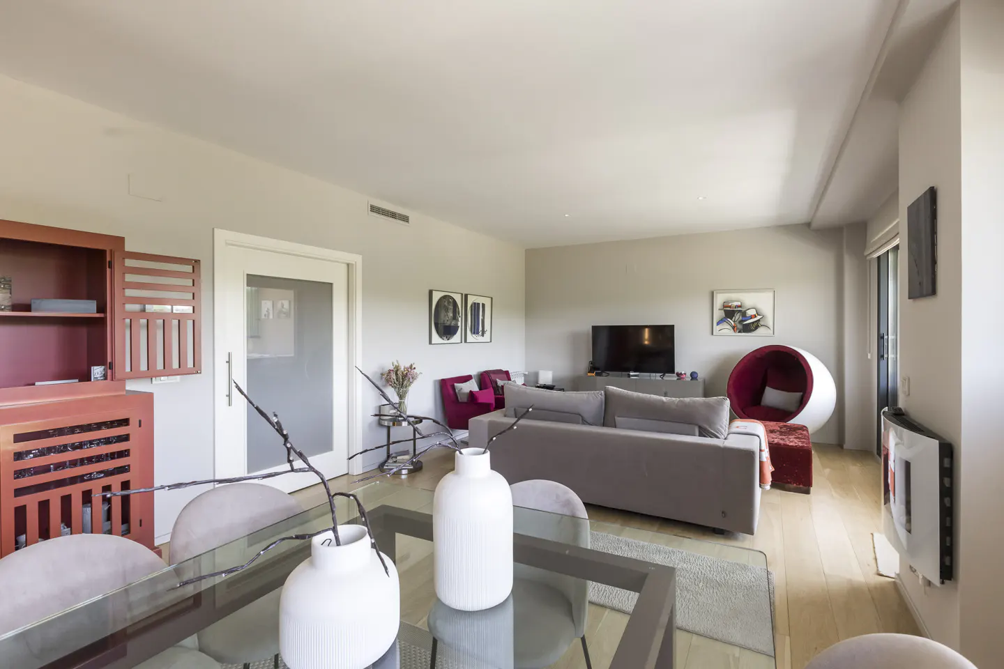 Modern living room with a glass dining table, grey sofa, red accent chairs, a unique egg chair, and a red storage cabinet.