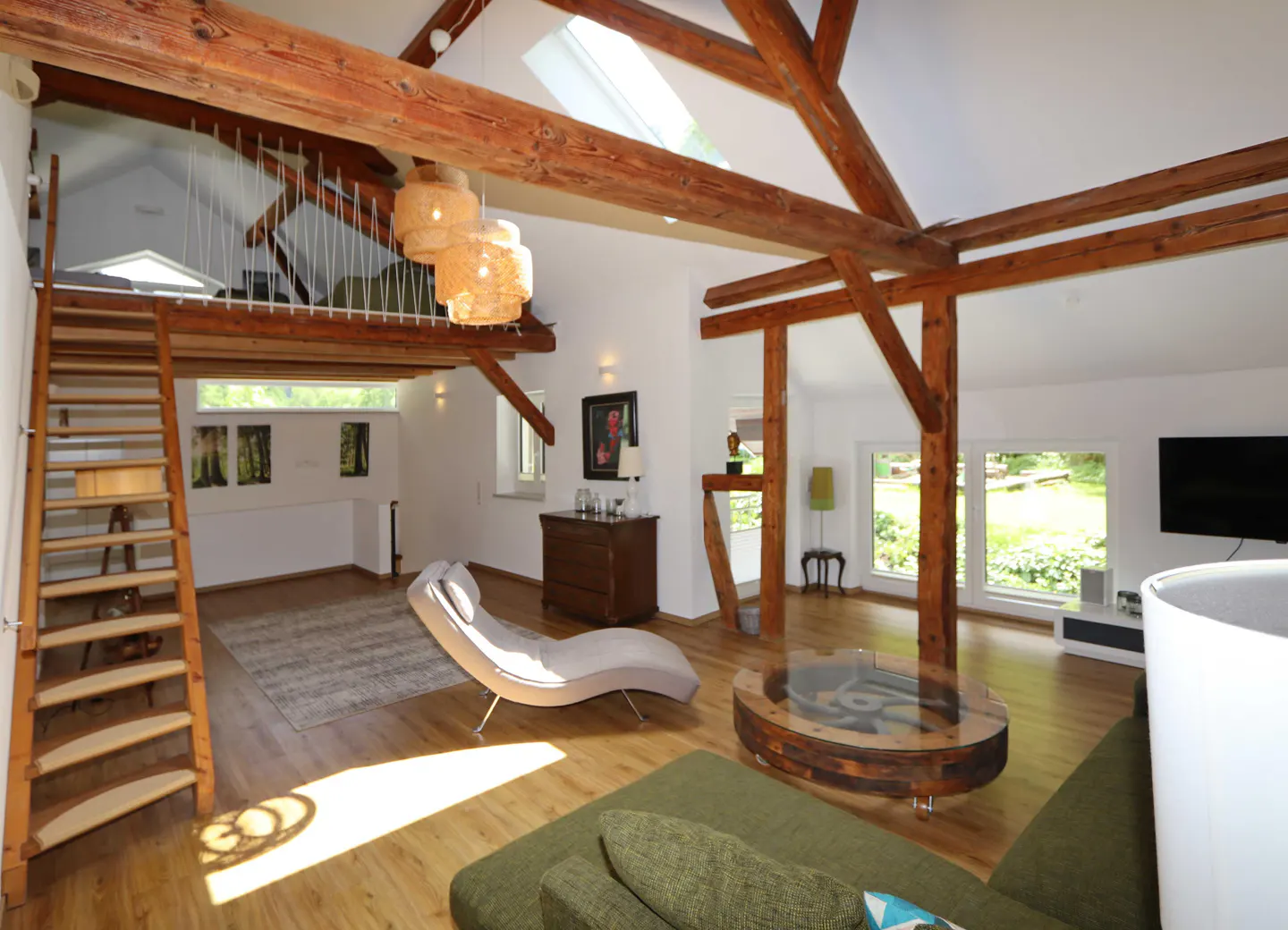 Bright living room with wood floors, exposed beams, and a loft with a wooden ladder. A chaise lounge and green sofa provide seating.