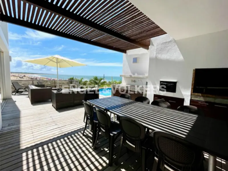 Outdoor patio with a long table, chairs, and lounge furniture overlooking the beach and ocean on a sunny day.