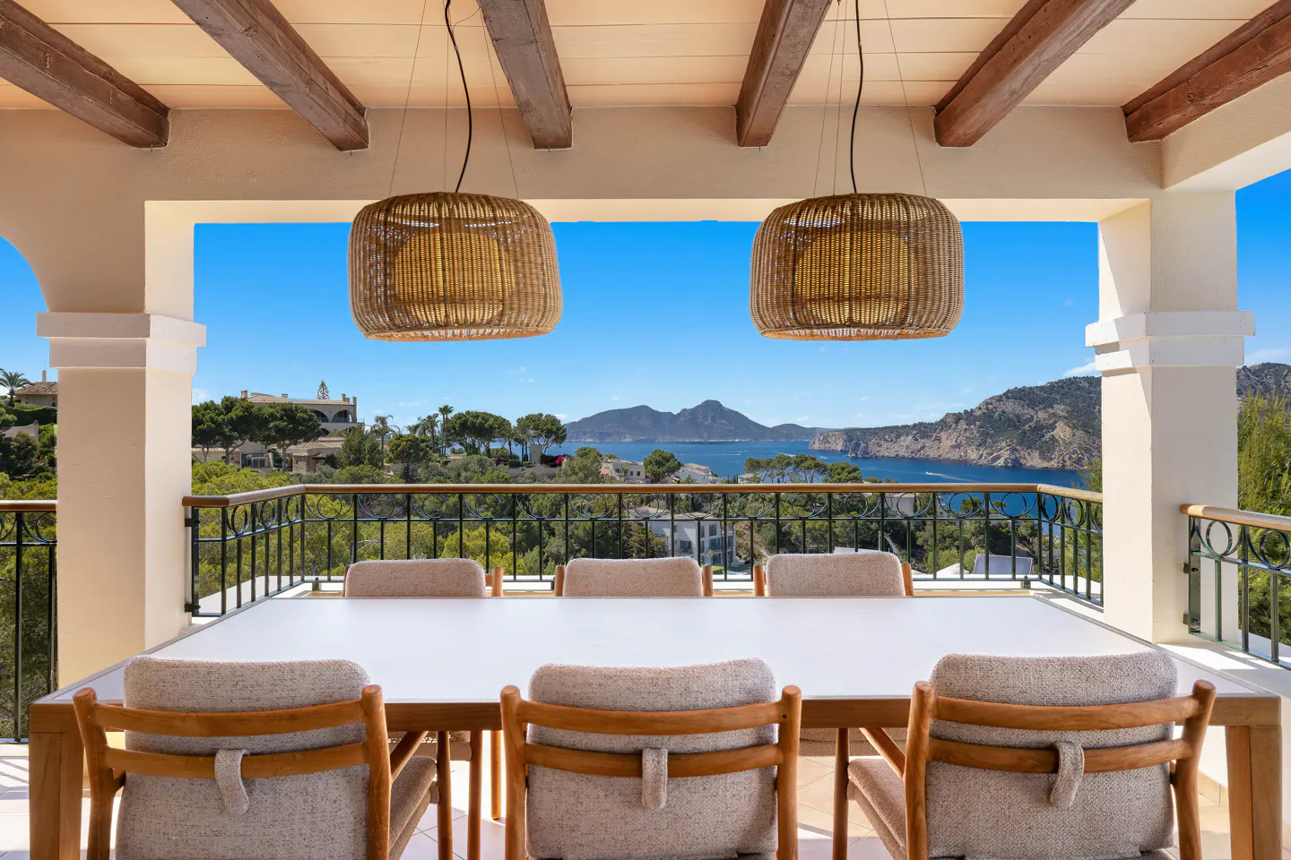 Outdoor dining area with a white table, chairs, and wicker pendant lights overlooking a scenic view of the ocean and mountains.