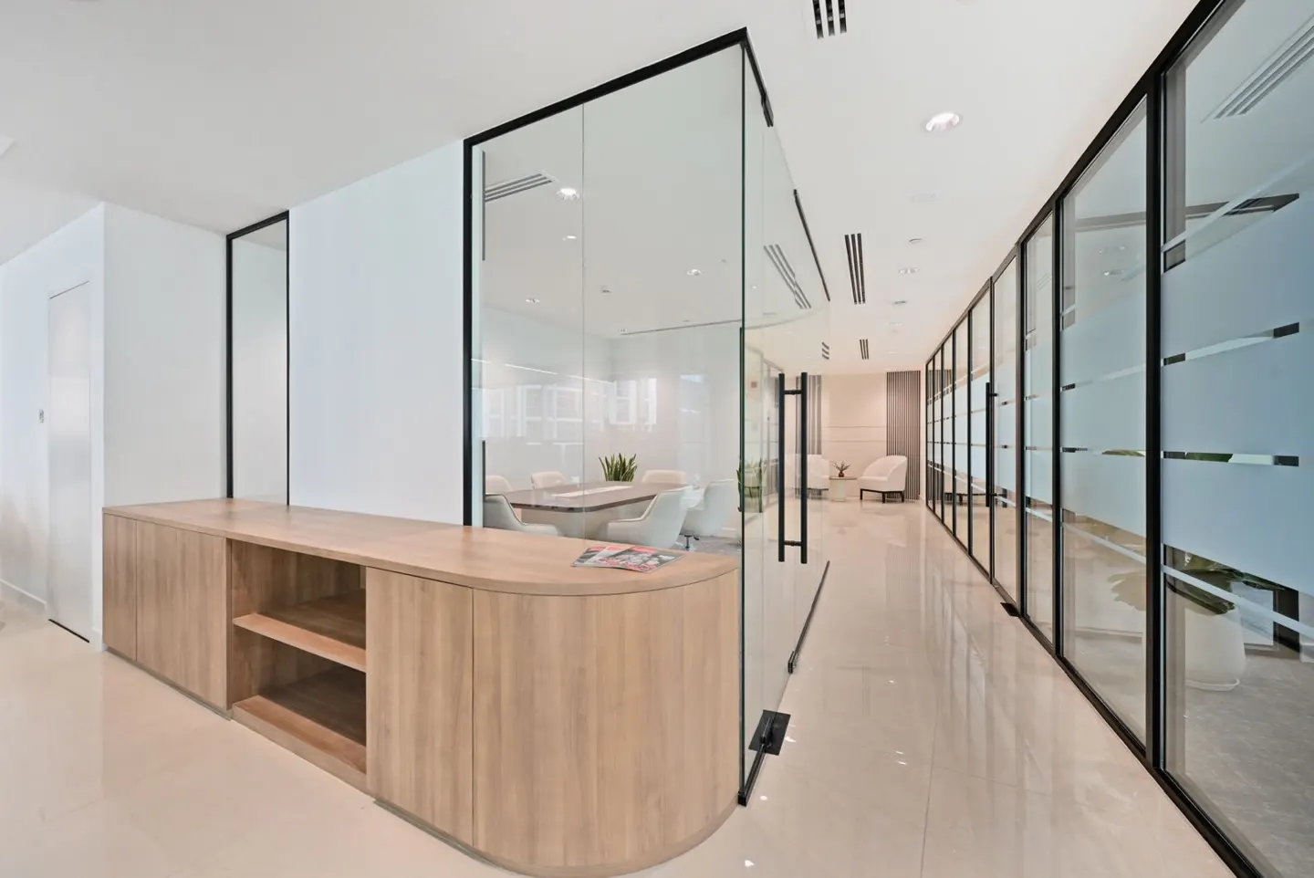 Bright office hallway with glass-walled rooms. A light wood cabinet is on the left. A conference room is visible through the glass.