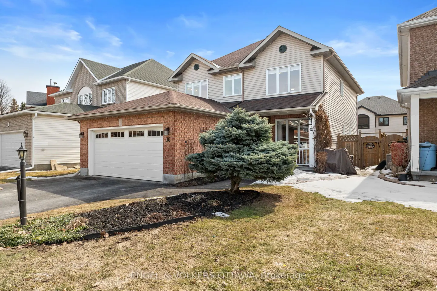 Two-story house with brick garage and beige siding under a blue sky. A small evergreen tree sits in the front yard.