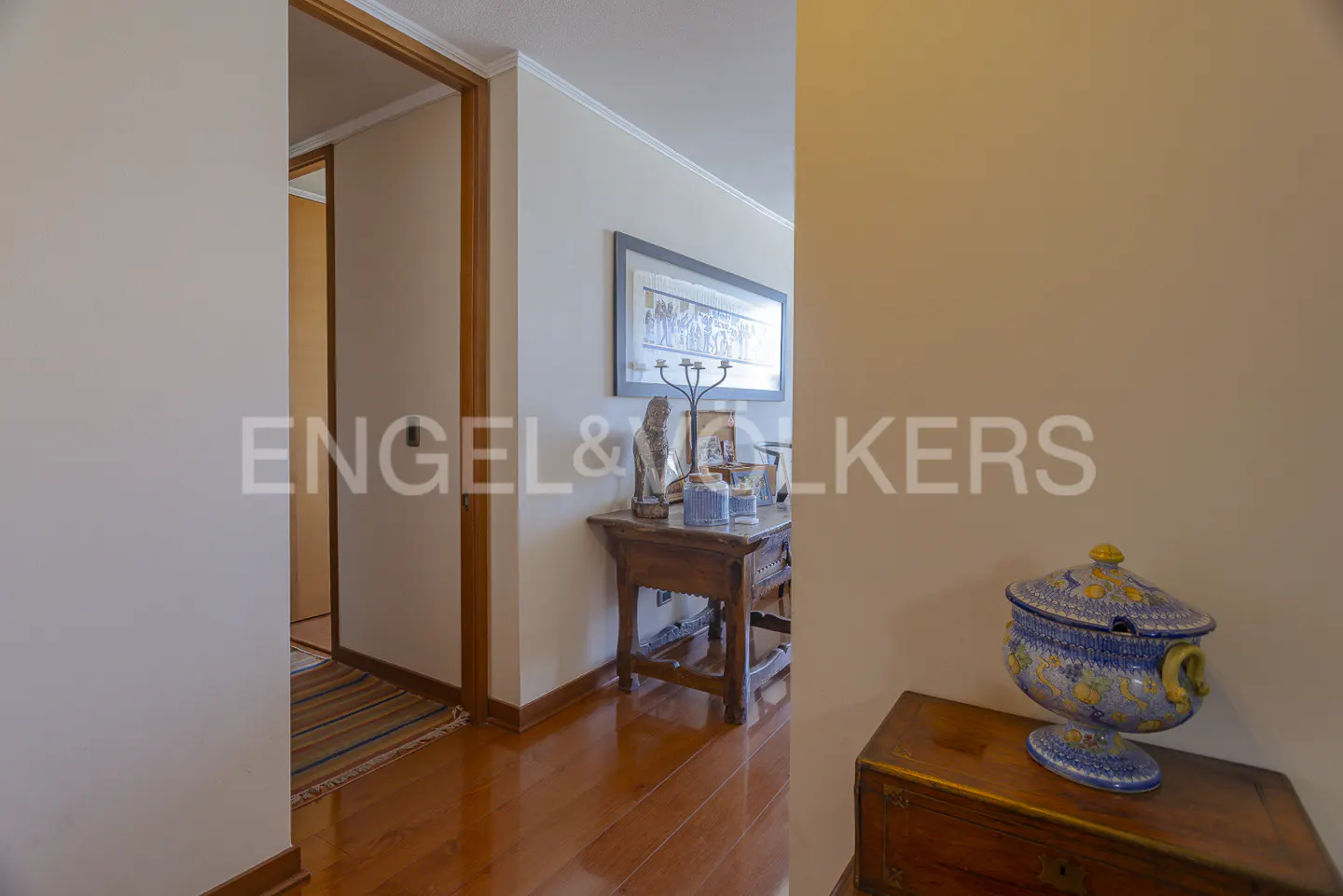 Hallway with wood floors, beige walls, and a blue decorative bowl on a wooden chest. A table with decor is visible.