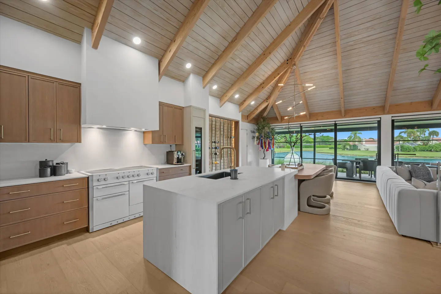 Open-concept kitchen with wood floors, white island, and vaulted wood ceiling. Large windows overlook a lake and outdoor seating.