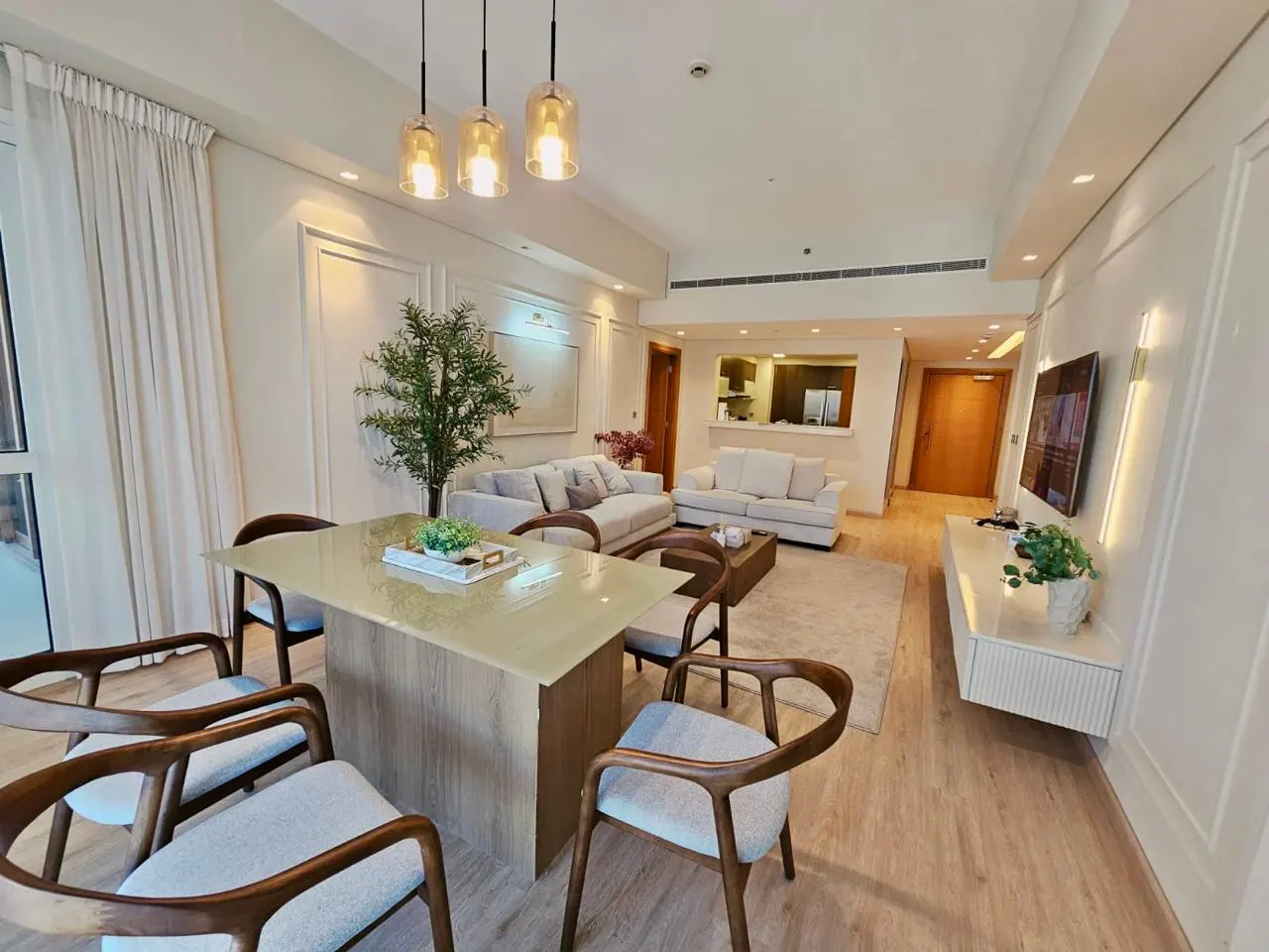 A bright, modern apartment interior with a dining table, sofas, and a TV. The color palette is neutral with wood accents.