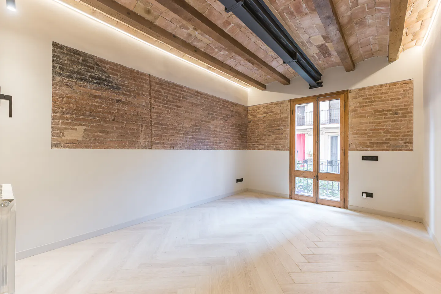 Empty room with herringbone wood floors, brick accent walls, and exposed wood beam ceiling. Double doors lead to a balcony.
