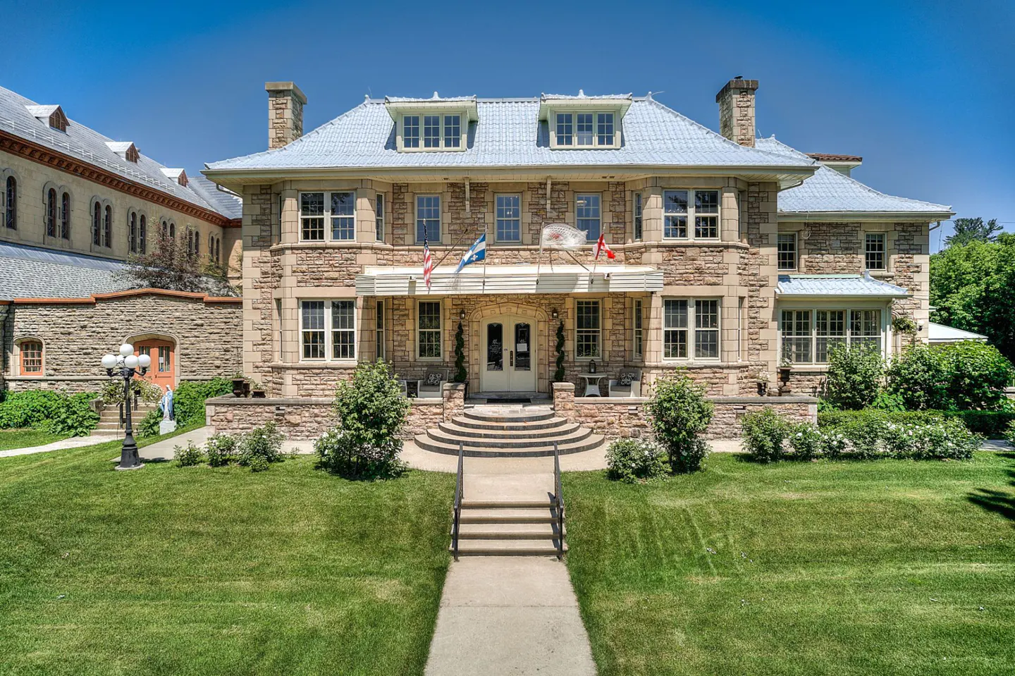 A large, two-story stone building with a blue tile roof and flags. A green lawn and walkway lead to the entrance.