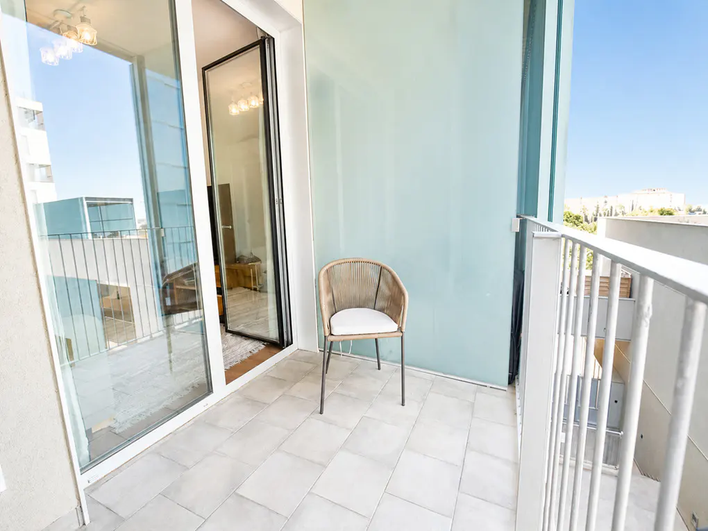 A bright balcony with a woven chair and white cushion. Sliding glass doors lead inside. White railings offer a city view under a blue sky.