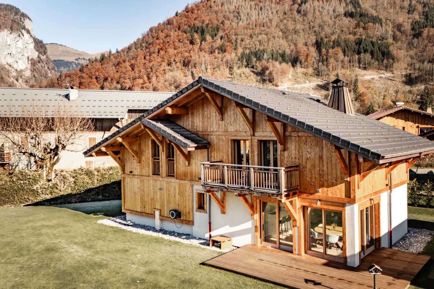 A two-story wood chalet with a balcony and a gray roof sits on a green lawn with mountains in the background.