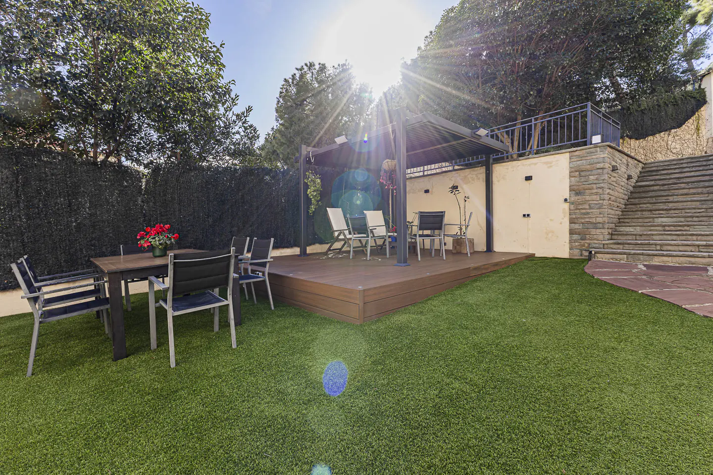 Backyard with green lawn, dining table with red flowers, and a wooden deck with a gazebo and chairs. Stone stairs on the right.