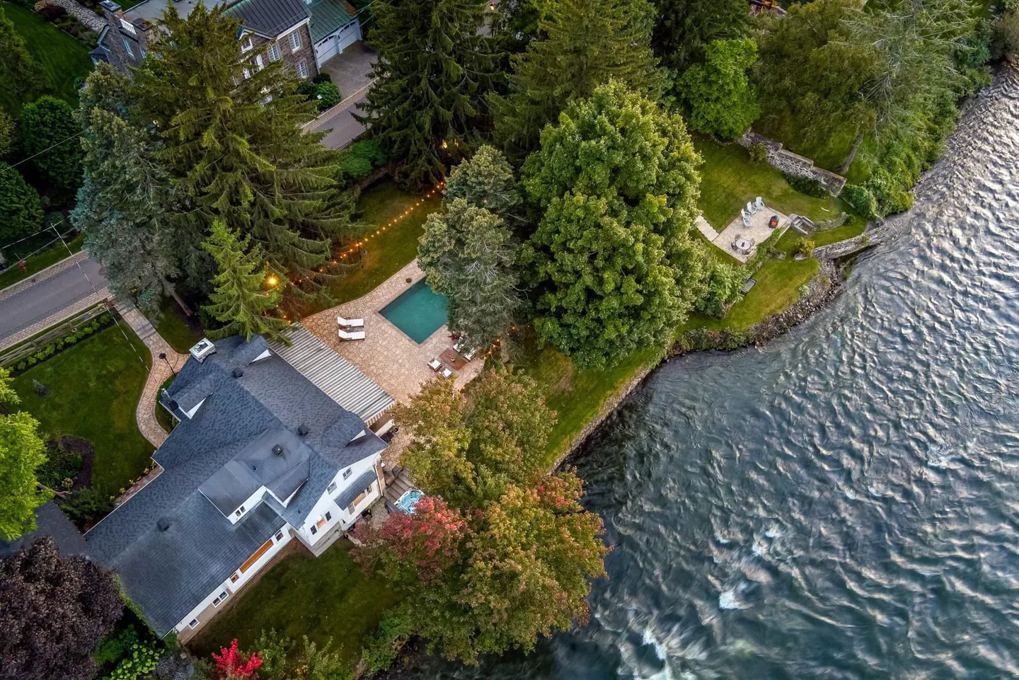 Aerial view of a white house with a dark roof, pool, patio, and riverfront access. Tall trees surround the property.